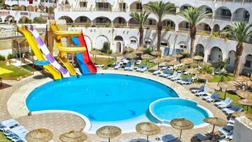 A pool area with slides, lounge chairs, umbrellas, and a building with balconies.