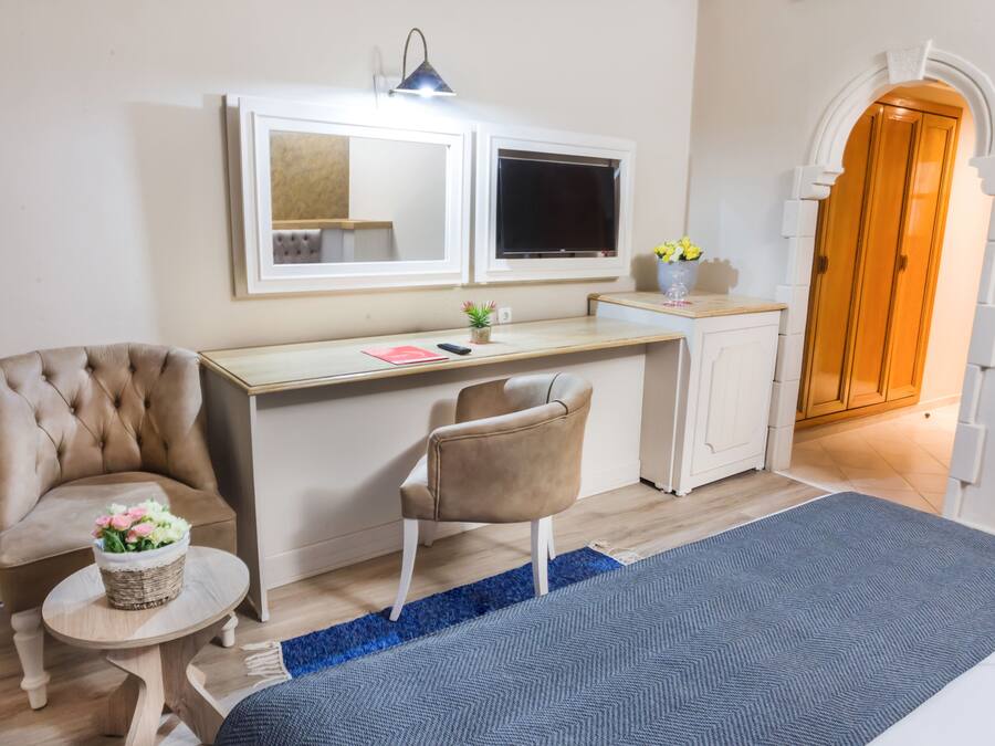Quarto de hotel com cama, escrivaninha com cadeira, mesinha com planta, espelho e guarda-roupa.