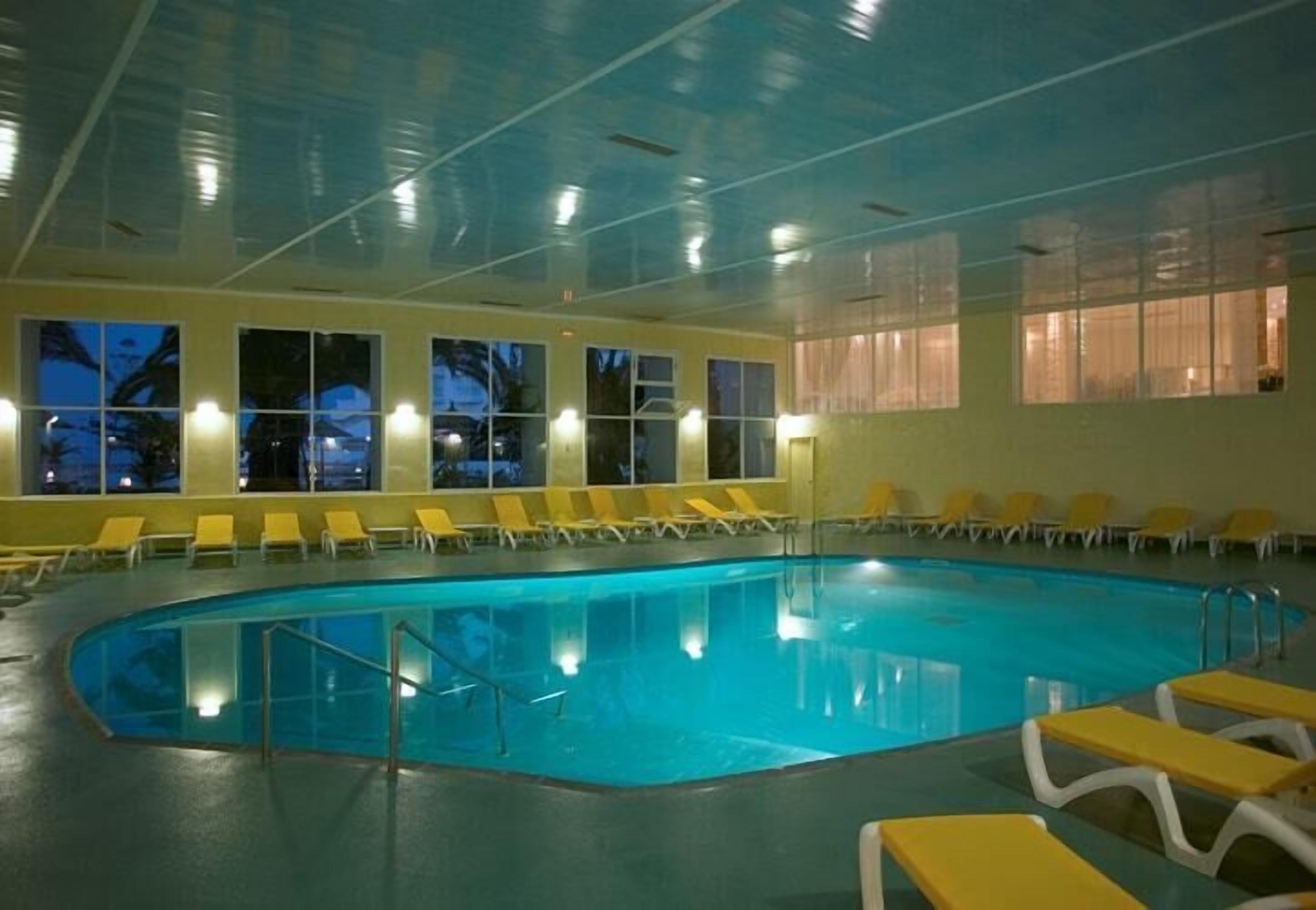 An indoor swimming pool with yellow lounge chairs and large windows.