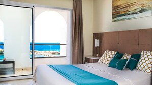 Un cuarto de hotel con cama, una mesita de noche, un ventanal con vista al mar y un cuadro en la pared.