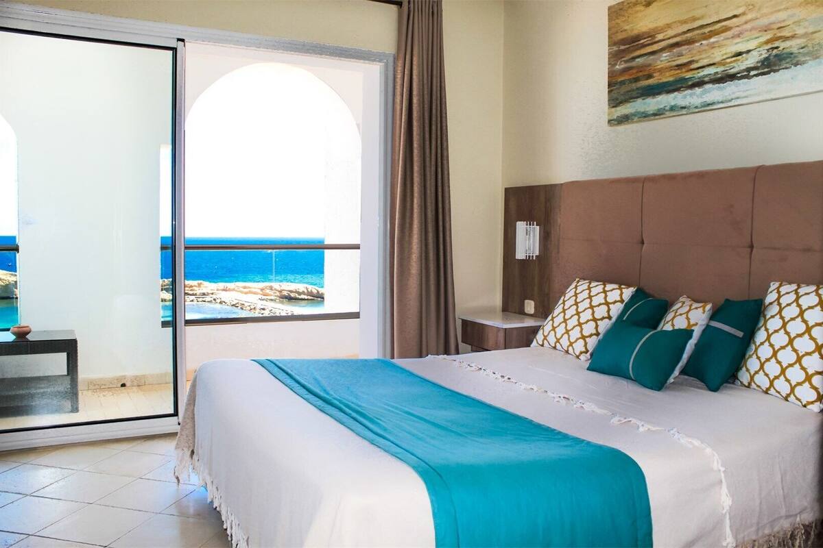 A hotel room with a bed, a nightstand, a window with a view of the sea, and a painting on the wall.