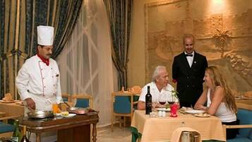 Comedor con un chef preparando comida, un camarero sirviendo y huéspedes sentados en una mesa.