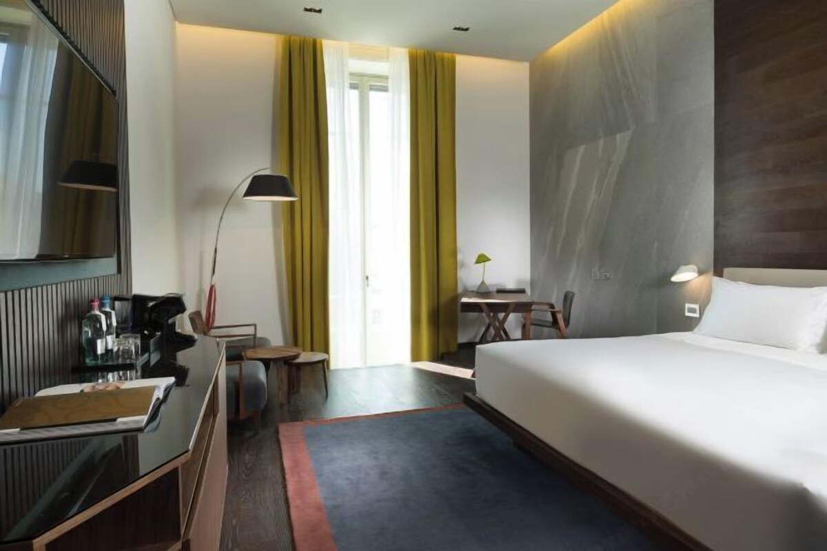 A hotel room with a bed, a desk with a chair, a lamp, and a window with curtains.