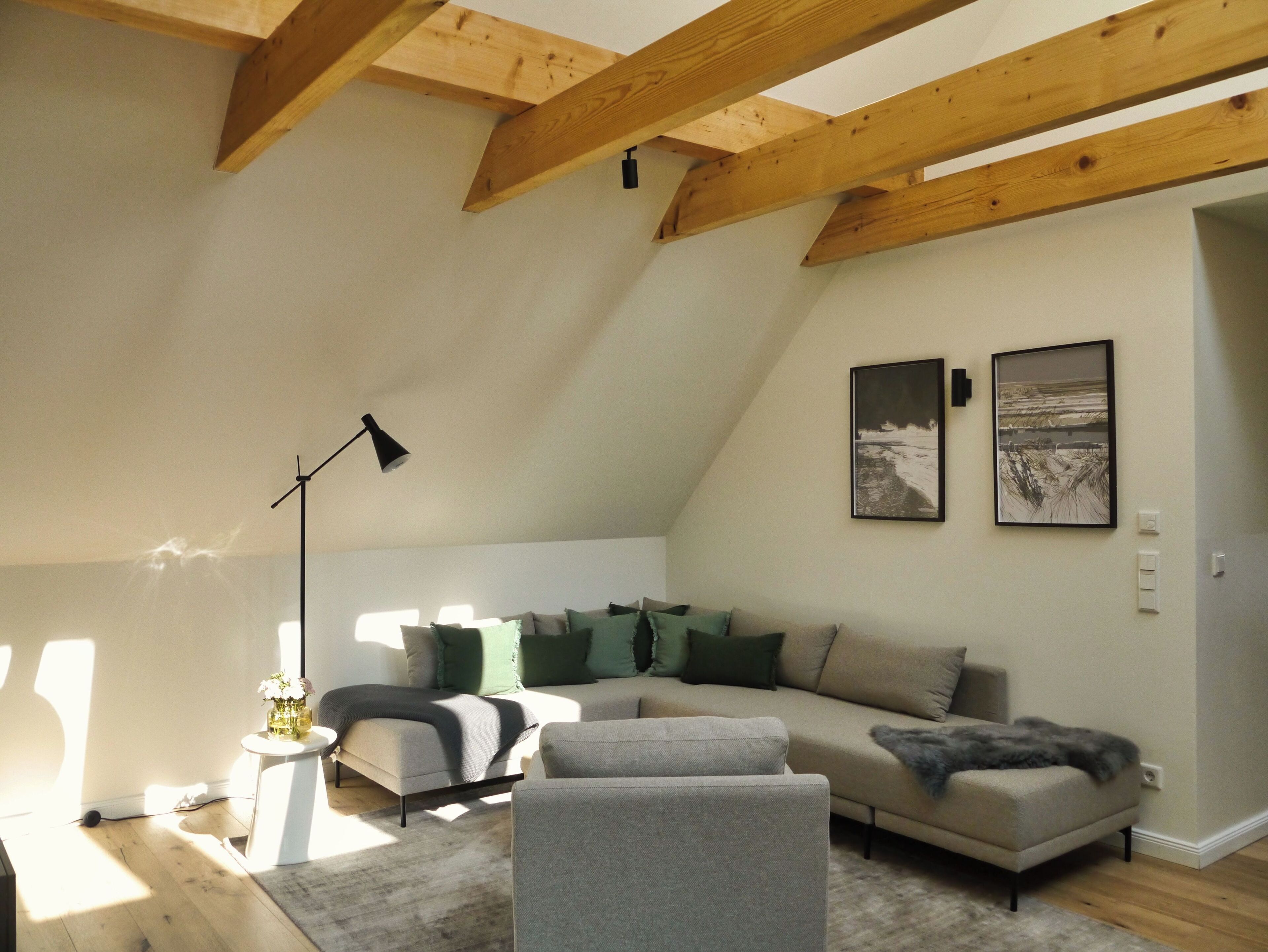 Une pièce chaleureuse de style loft, avec un plafond en pente, des poutres apparentes, un canapé d’angle, un petit fauteuil, un lampadaire et deux tableaux encadrés accrochés au mur.