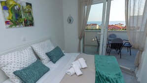 Une chambre à coucher avec un lit, des oreillers, une couverture verte, un tableau et une vue sur la mer depuis un balcon.