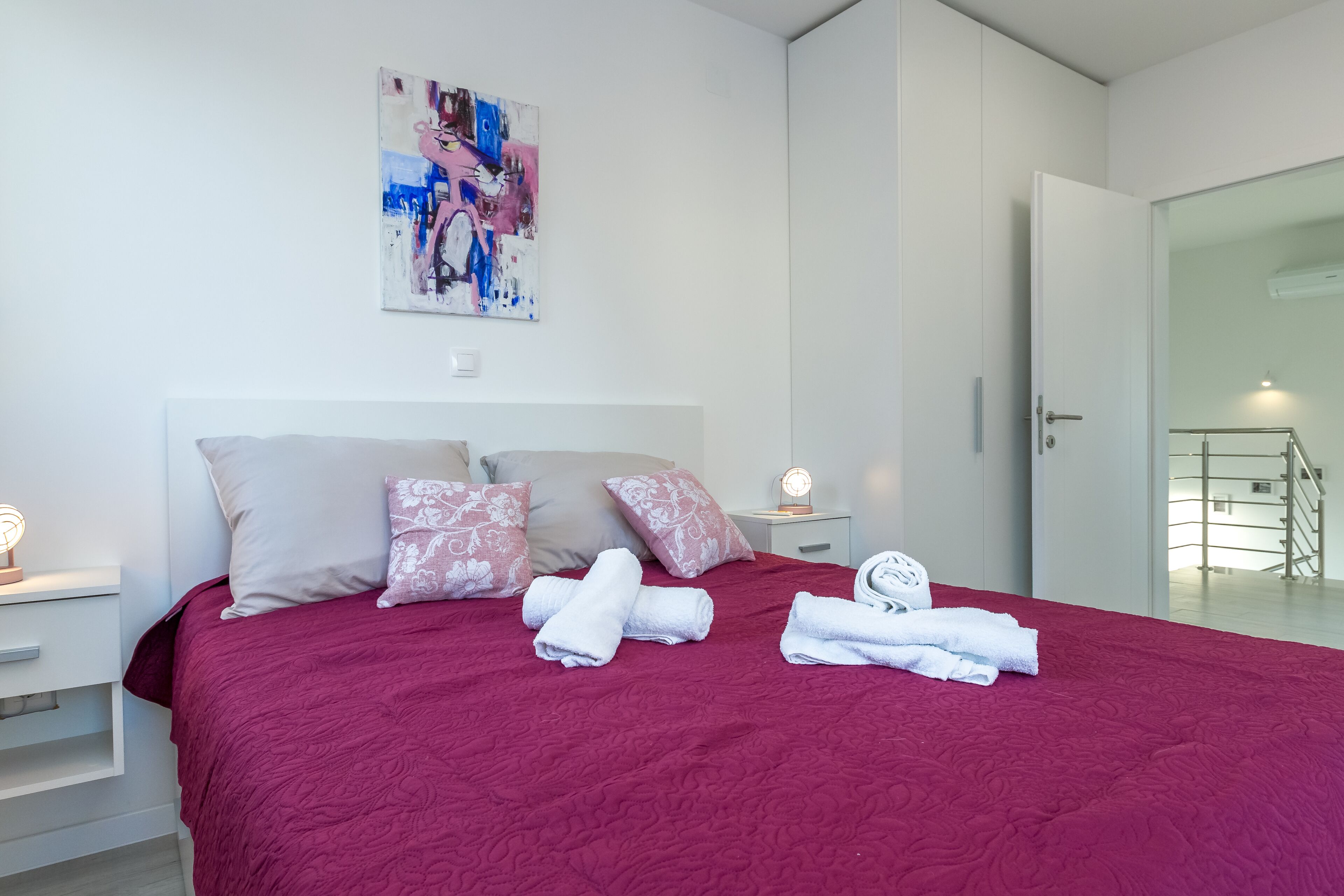 Una cama bien hecha con una colcha roja, almohadas blancas y toallas enrolladas. Hay un cuadro en la pared encima de la cama y una puerta que da a una escalera.