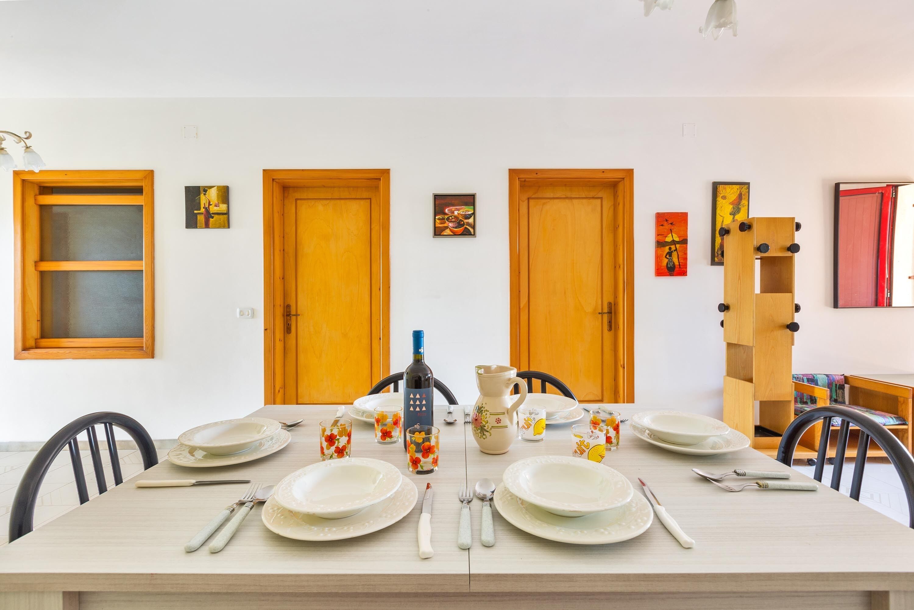 Mesa de jantar posta com pratos, talheres e copos, ao lado de portas de madeira e obras de arte emolduradas.