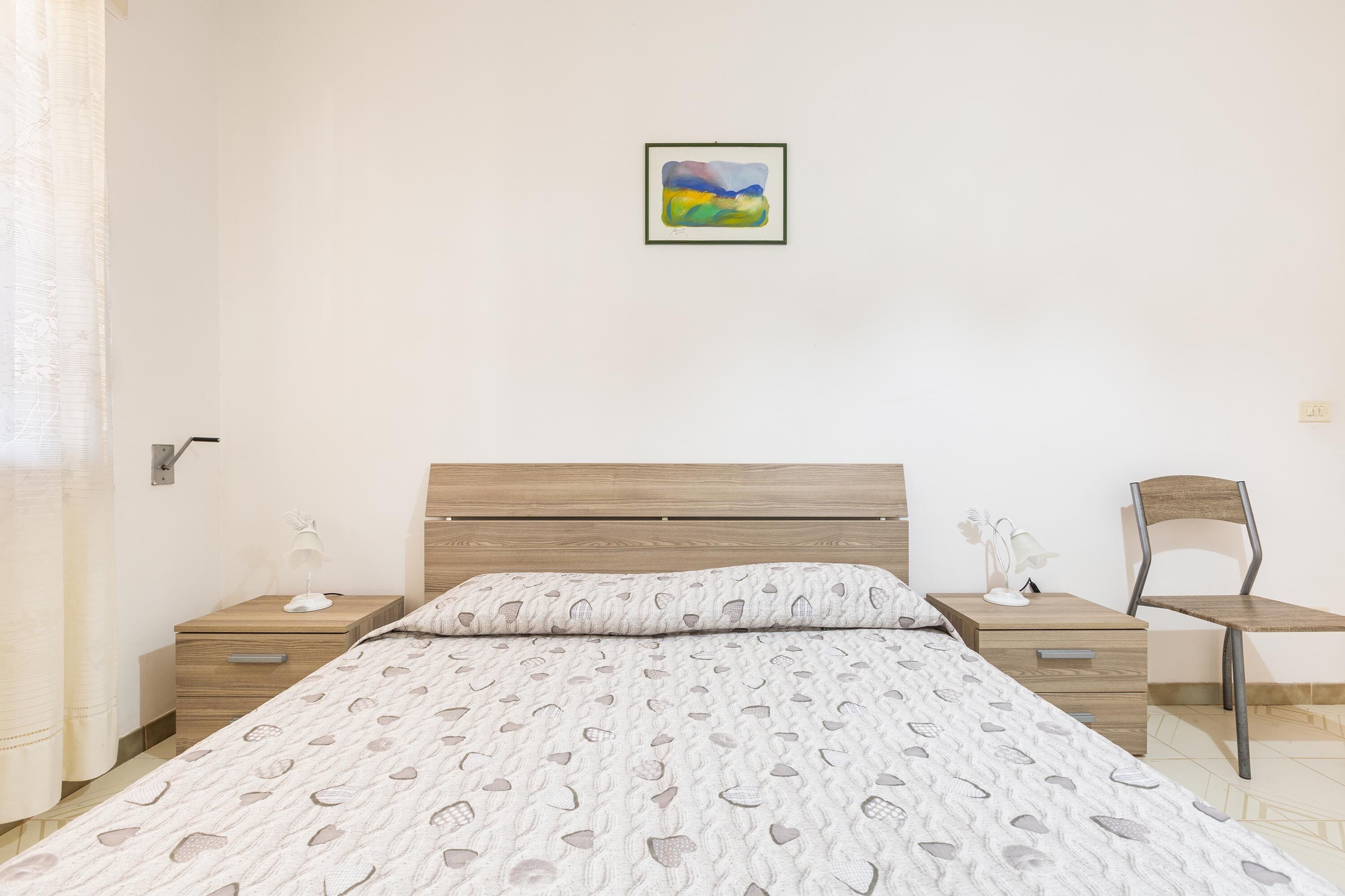 A bedroom with a bed, a nightstand, a chair, and a wall-mounted painting.