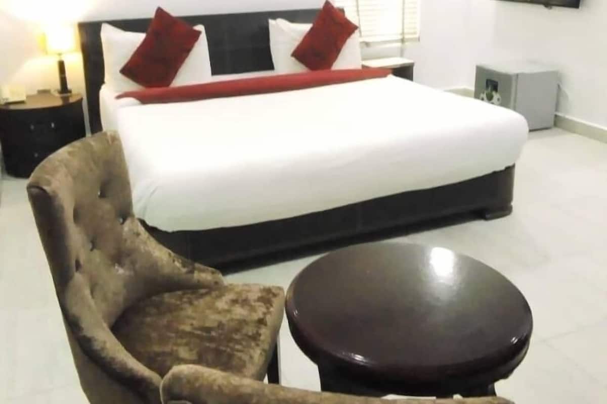 Ein Hotelzimmer mit Bett, Sessel, kleinem Tisch, Klimaanlage, Fernseher und Fenster mit Jalousien.