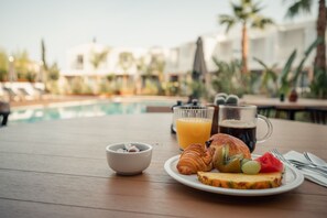 A breakfast setting with croissants, fruit, a coffee mug, and a glass of orange juice on a wooden table by a poolside. - Van der Valk Hotel Bloom Saint Aygulf (Fréjus)