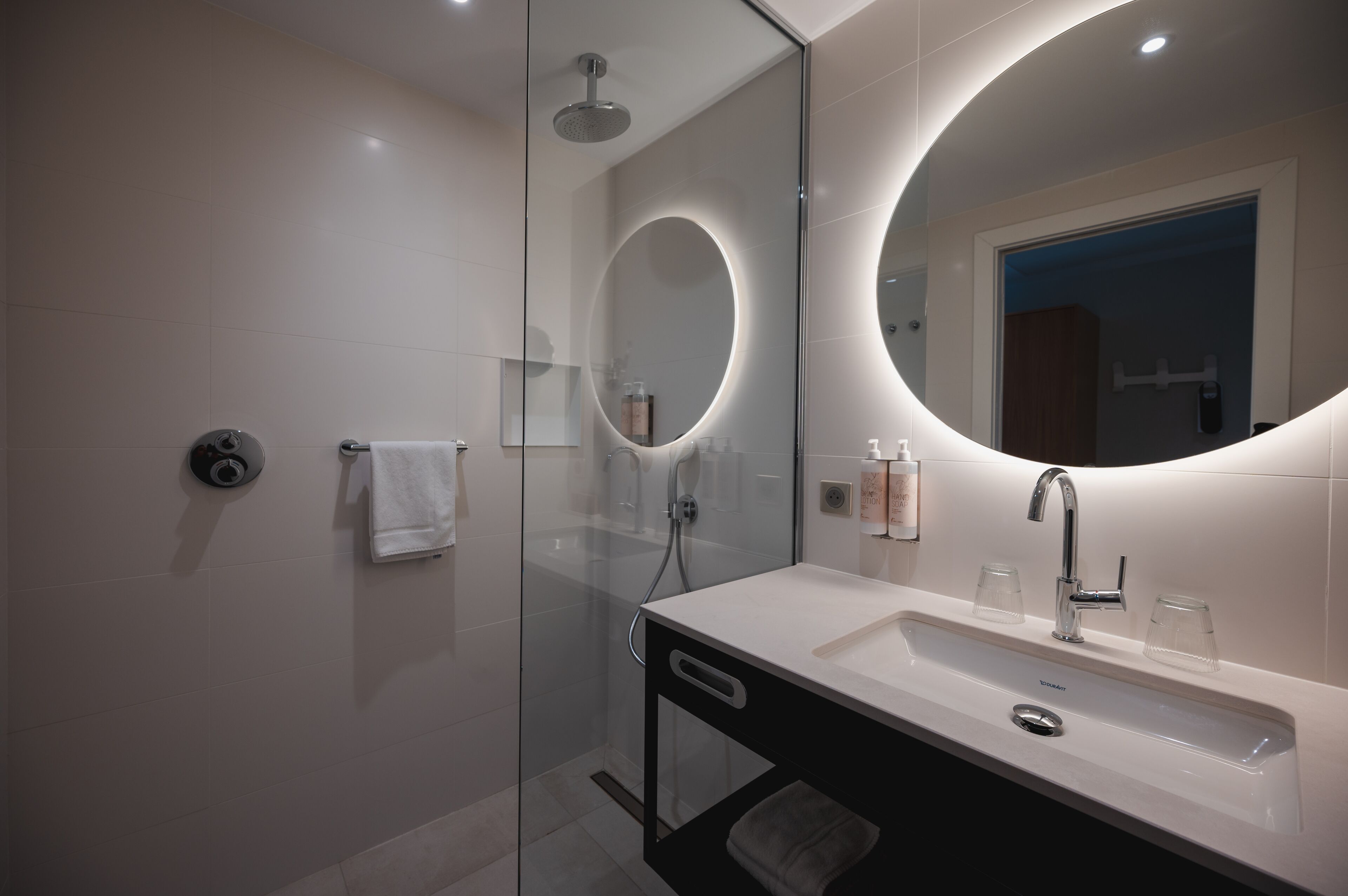 Un baño moderno con ducha de vidrio, un lavamanos blanco con espejo y dos vasos transparentes sobre el lavamanos.