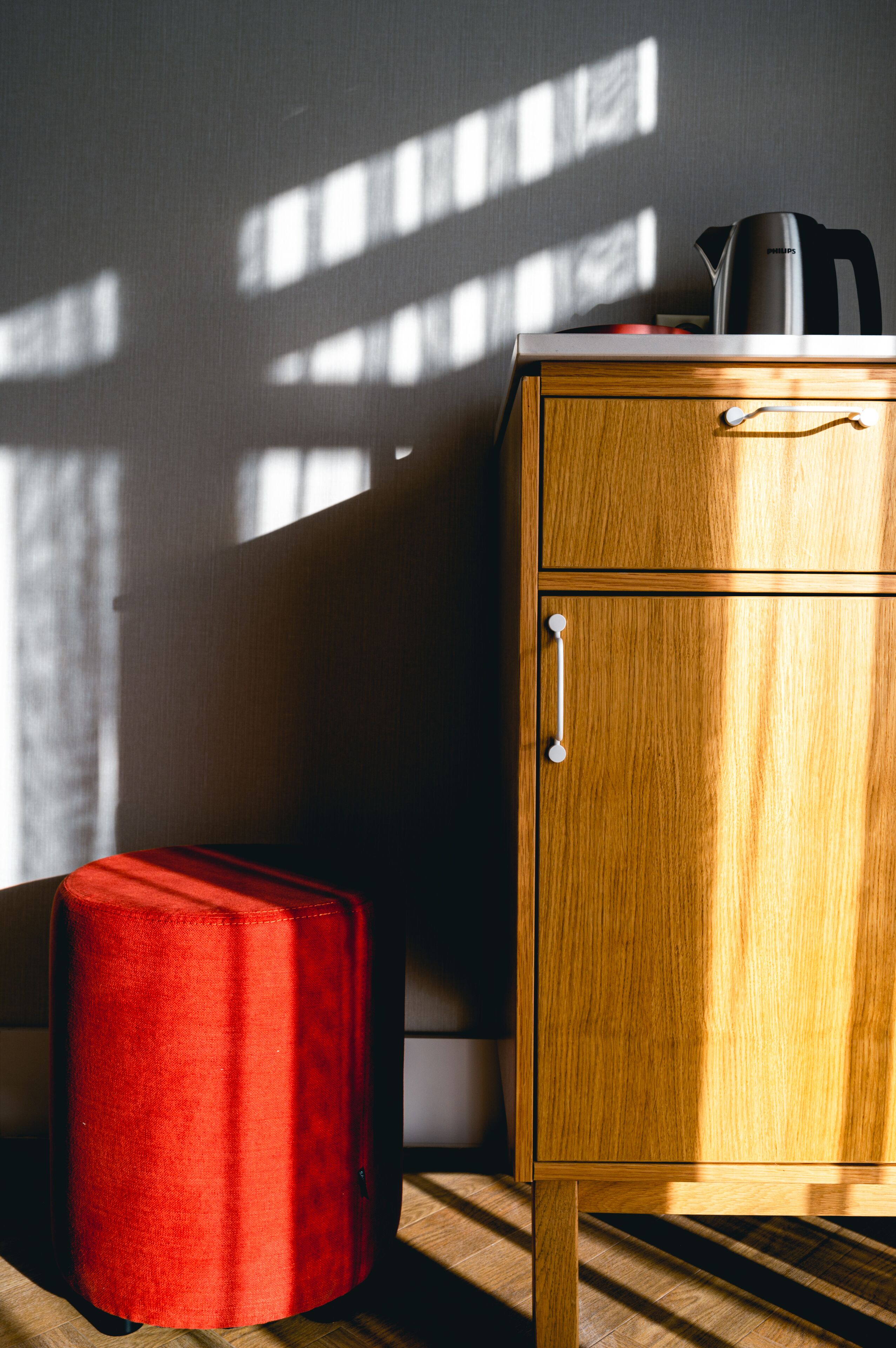 Un meuble en bois sur lequel est posée une bouilloire, un pouf rouge et une fenêtre qui projette des ombres sur le mur.