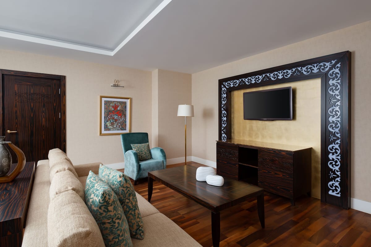 A living room with a sofa, a teal armchair, a coffee table, a TV mounted on a decorative wall, and a wooden door.