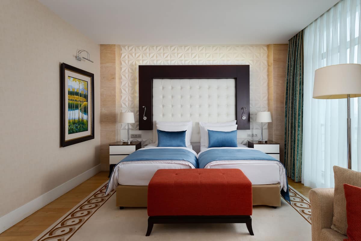 A hotel room with a large bed, two bedside tables, a nightstand, a red bench, a floor lamp, and a painting on the wall.
