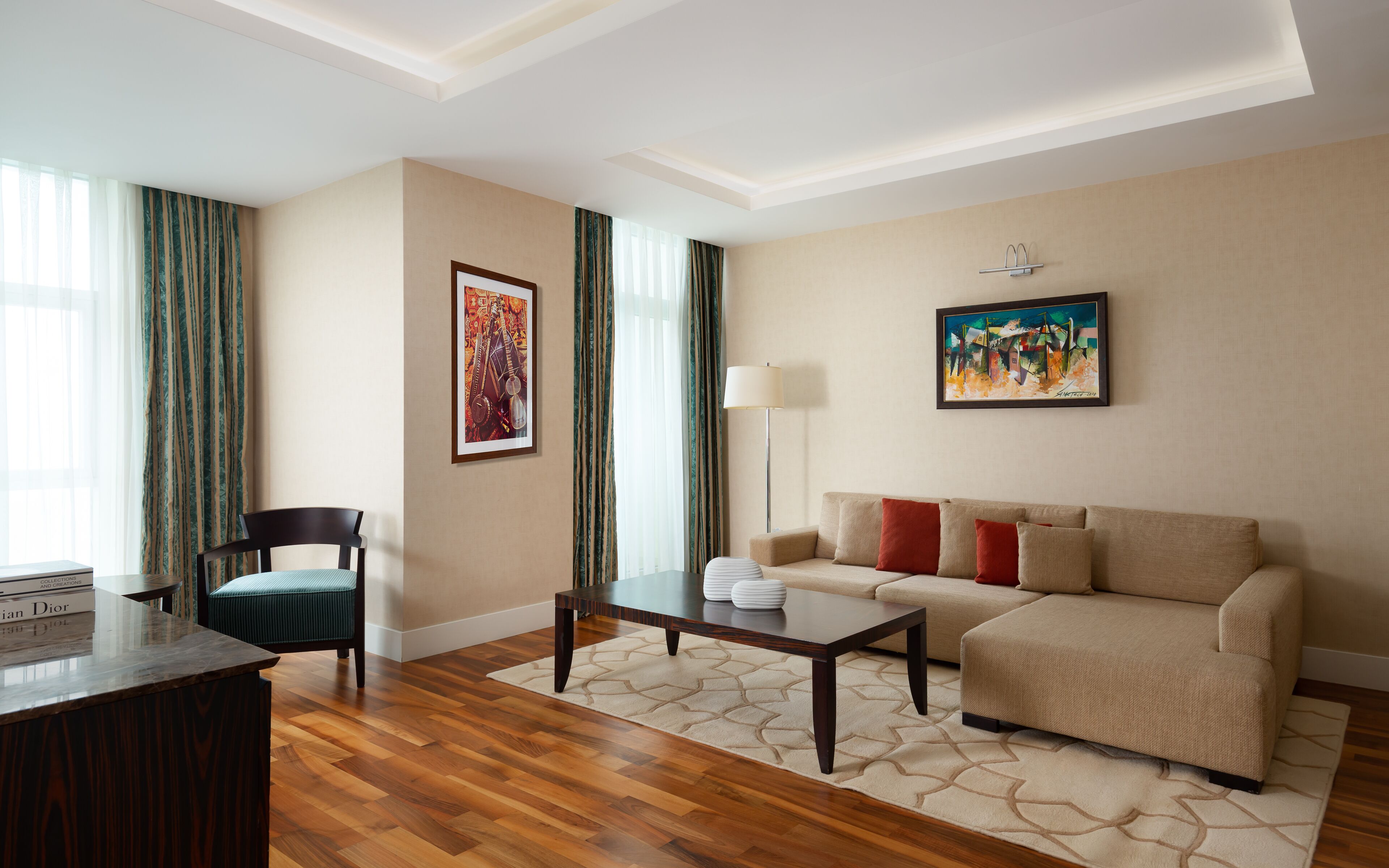 Habitación de hotel con suelo de madera, un sofá, una silla, una mesa de centro, un escritorio y dos cuadros en las paredes.