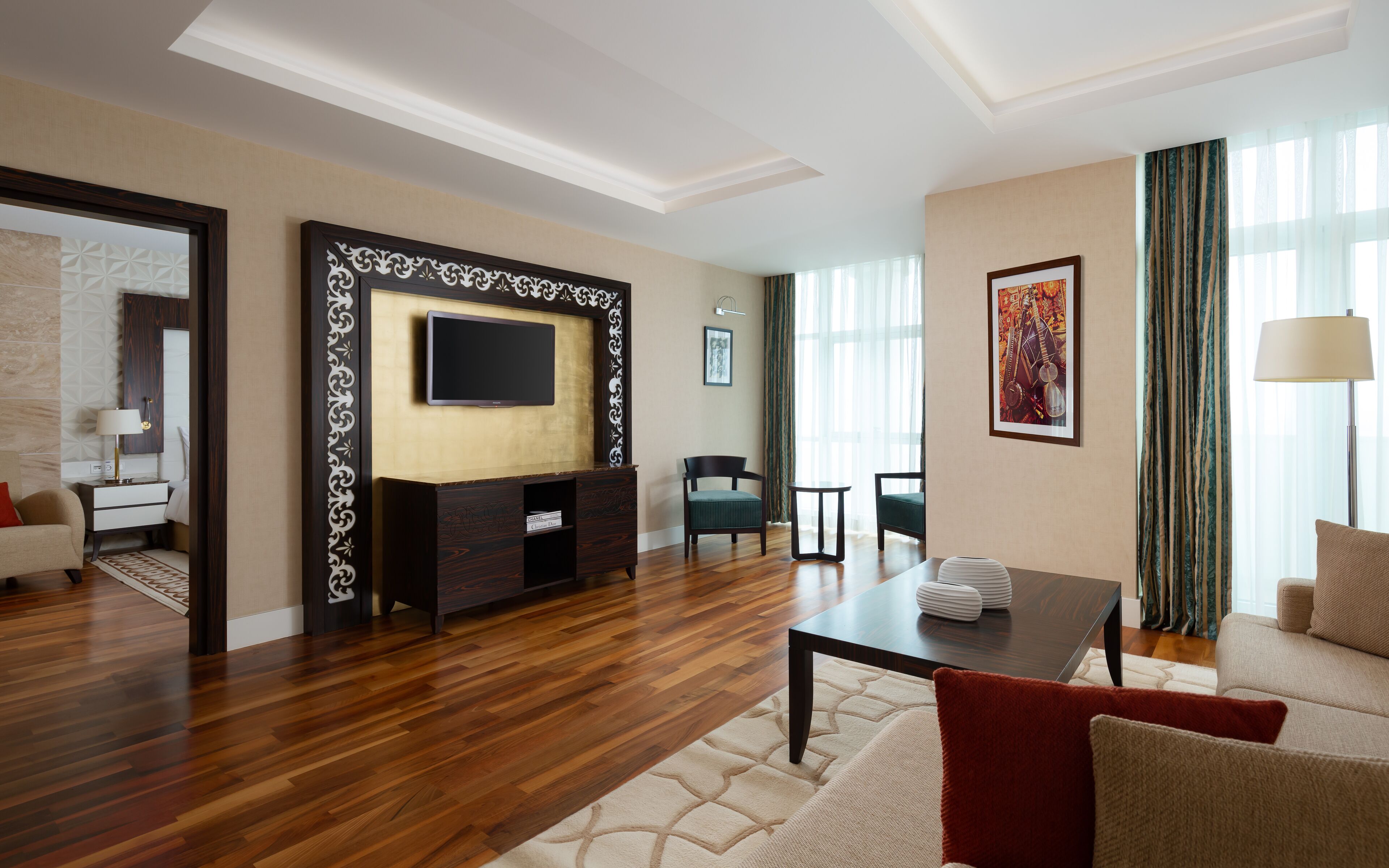 A modern hotel room with a wooden floor, a flat-screen TV mounted on a dark wooden wall, a desk with a chair, a dining table with chairs, and a large window with sheer curtains.