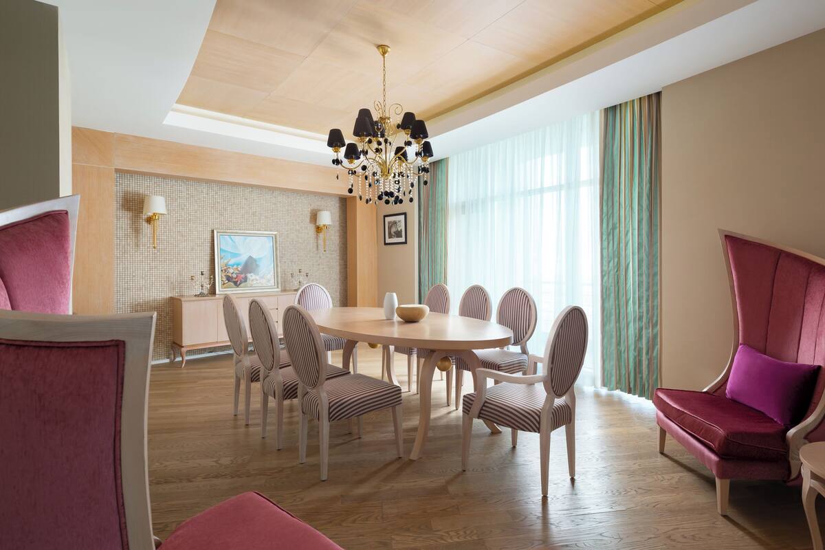 A dining area with a round table, striped chairs, a chandelier, and a view of a green curtain.