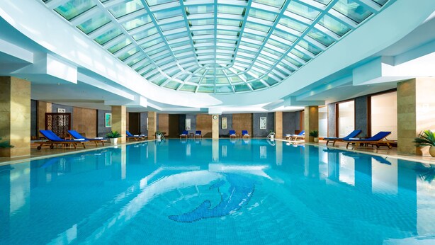Una piscina interior con agua azul cristalina, rodeada de sillones de descanso y un techo de vidrio.