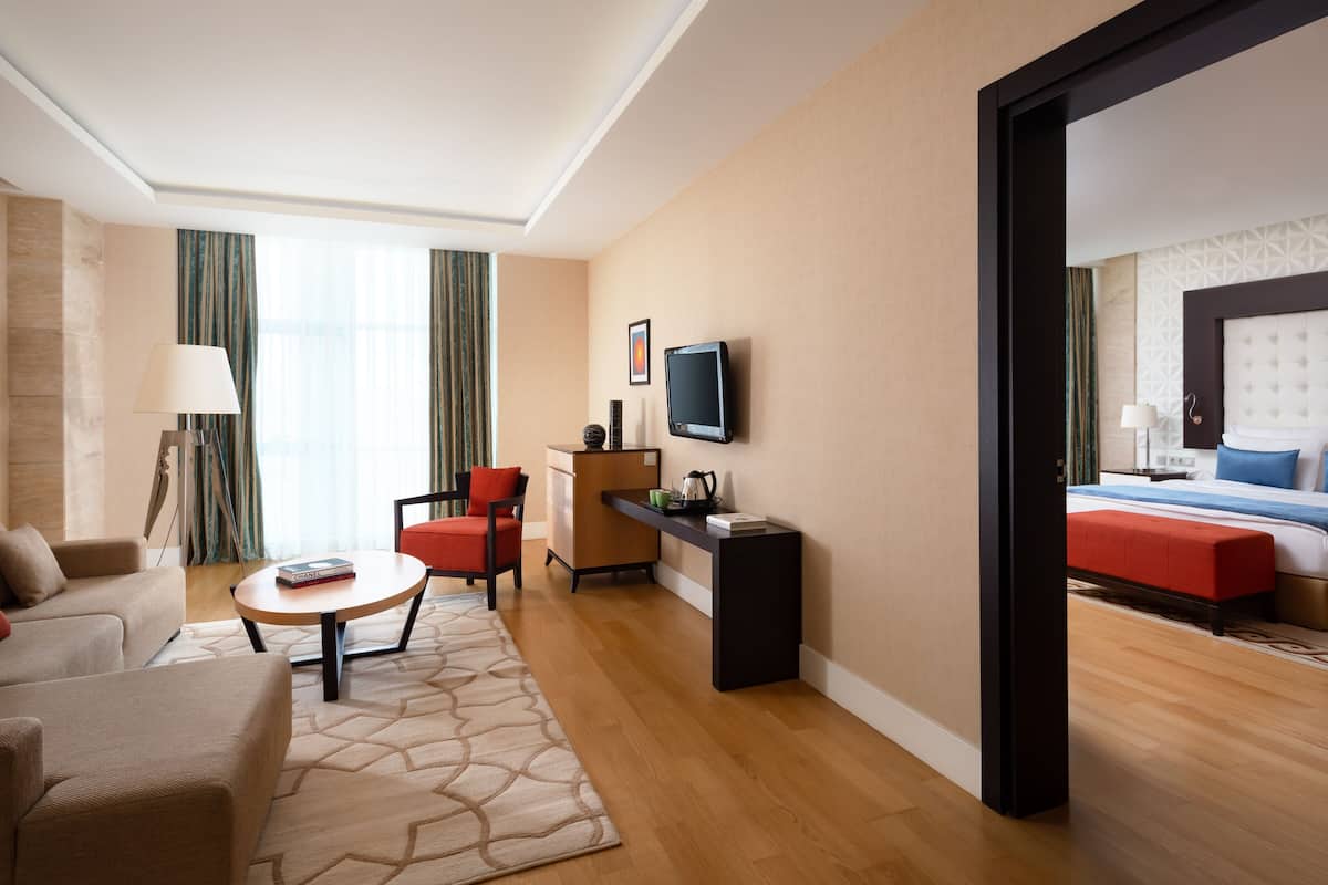 A hotel room with a sofa, armchair, coffee table, TV, and a bed with a red accent.