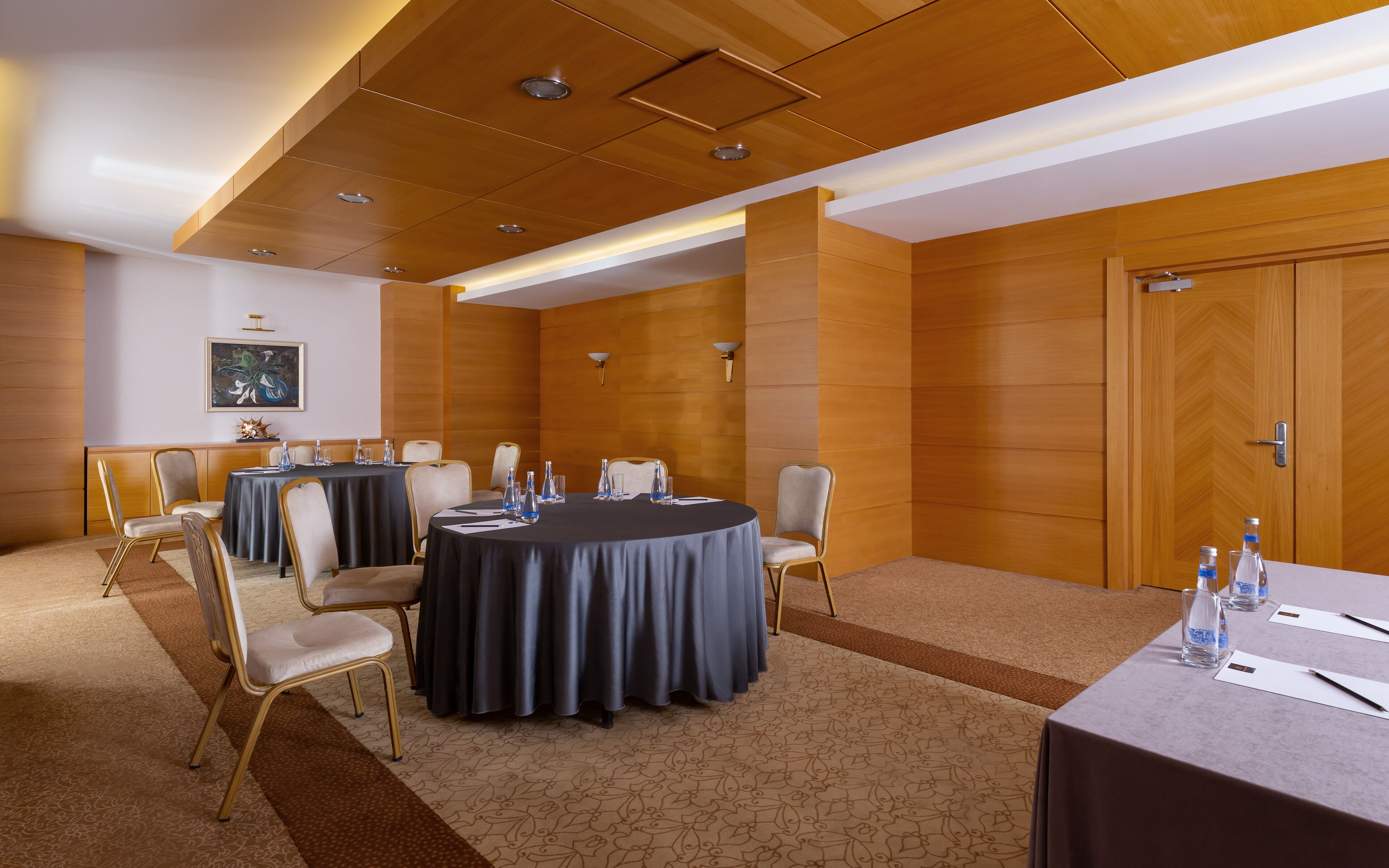 Un salón de conferencias con mesas redondas, sillas y una pared de madera con un cuadro.