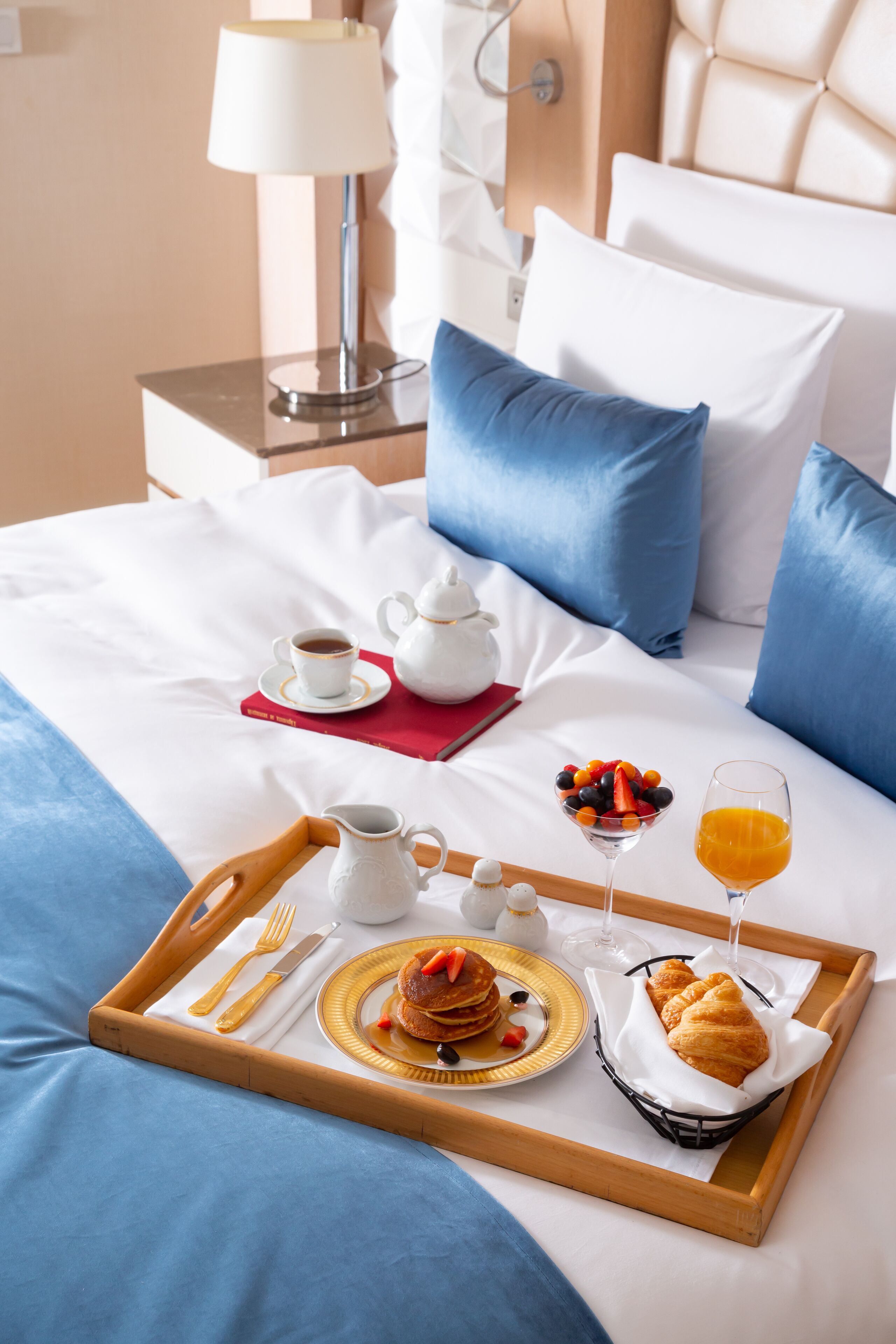 A neatly made breakfast tray with pancakes, a croissant, fruit, and orange juice on a bed with blue bedding and white pillows.