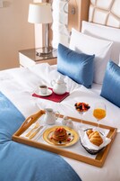 Una bandeja de desayuno bien presentada con tortitas, un croissant, fruta y zumo de naranja sobre una cama con ropa de cama azul y almohadas blancas.