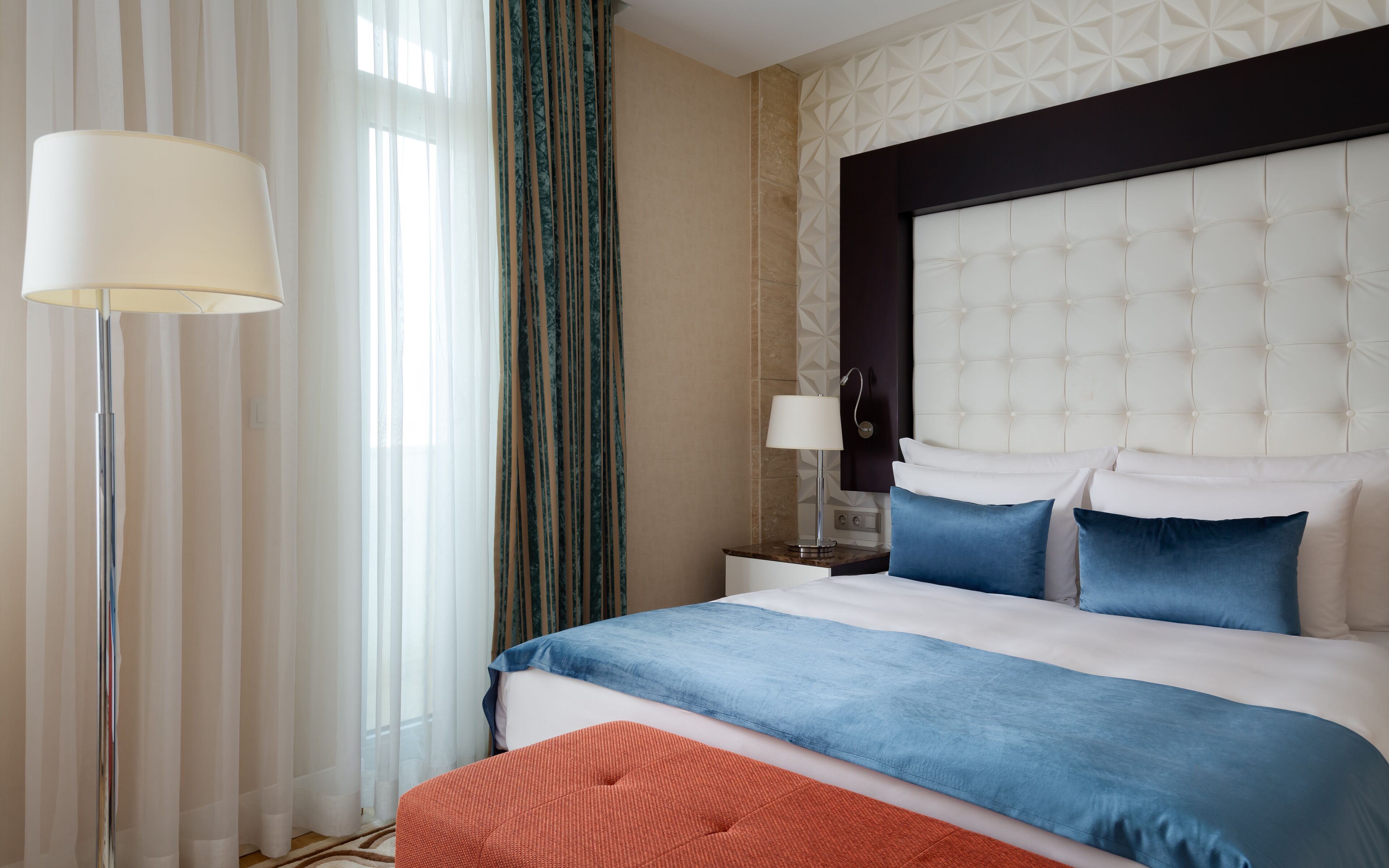 A hotel room with a large bed, a nightstand, a lamp, and a view of the outside through sheer curtains.