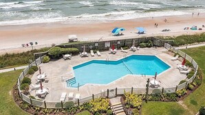 A swimming pool with a deck area, surrounded by a fence, and overlooking a beach with people and umbrellas. - 3BR Oceanfront Condo with Community Pool (North Topsail Beach)