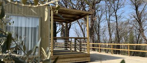 A wooden cabin with a covered porch and a fenced deck, surrounded by trees and gravel.
