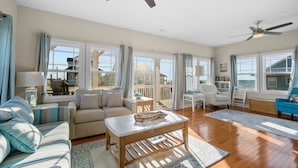 A spacious living room with a sofa, coffee table, and chairs, large windows with curtains, and ceiling fans.