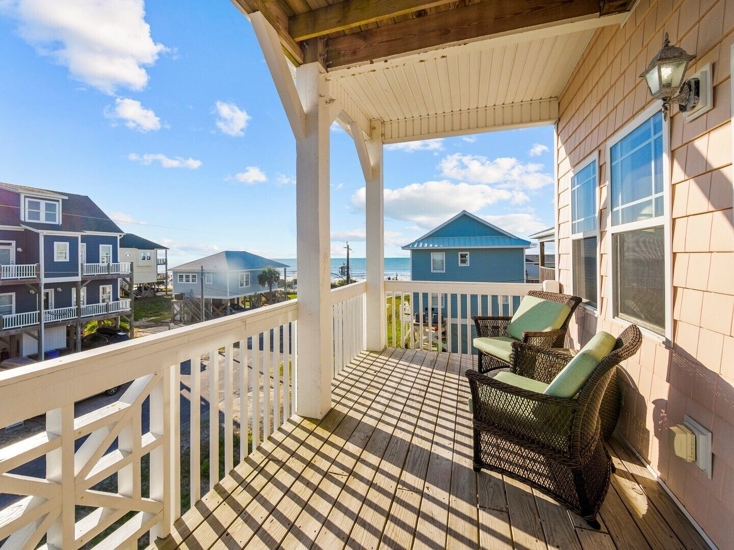 A wooden deck with a white railing, wicker chairs, and a view of neighboring houses and the ocean.
