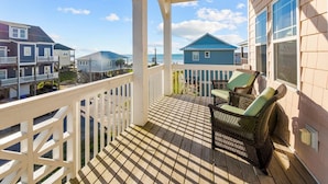 A wooden deck with a white railing, wicker chairs, and a view of neighboring houses and the ocean.