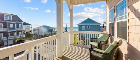 A wooden deck with a white railing, wicker chairs, and a view of neighboring houses and the ocean.