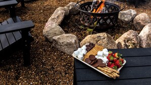 Un montaje para picnic con una bandeja de aperitivos que incluye malvaviscos, chocolate y fresas, rodeado de rocas y un brasero.