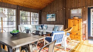 Interior acogedor de una cabaña con techo de madera, una mesa de comedor con sillas, un sofá con cojines, un sillón de mimbre y un armario.
