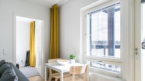 A modern living room with a sofa, a dining table with chairs, a small plant, and a window with a view. - Hiisi Homes Kirkkonummi Laajakallio (Kirkkonummi)
