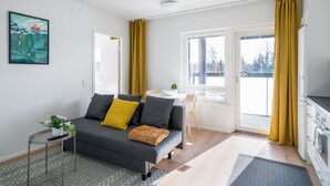 A modern living room with a grey sofa, yellow cushions, a small side table with a plant, and a kitchen area with a stove and cabinets. - Hiisi Homes Kirkkonummi Laajakallio (Kirkkonummi)
