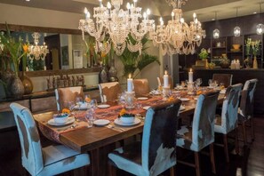 A dining area with a large wooden table set for a meal, surrounded by chairs with blue upholstery. A grand chandelier hangs above the table, and the room features dark wood paneling and a large mirror on the wall.