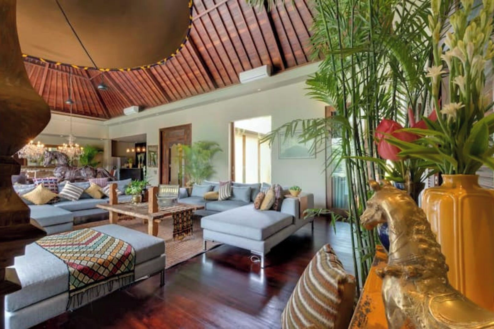 A spacious living area with a high ceiling, a large sofa set, and a coffee table. There are potted plants and a decorative vase on a side table.