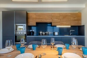 Cozinha moderna com armários de madeira, eletrodomésticos de aço inoxidável e área de refeições para seis pessoas, com louças azuis.