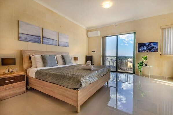 A modern bedroom with a large bed, a nightstand, a TV, and a balcony with a view.