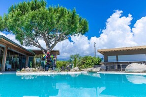 Zona de piscina con un árbol grande, un espacio de estar cubierto y un edificio con ventanales.