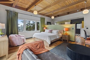 A modern bedroom with a large bed, a sitting area with a chair, a small table, and a view of the outdoors through the window.