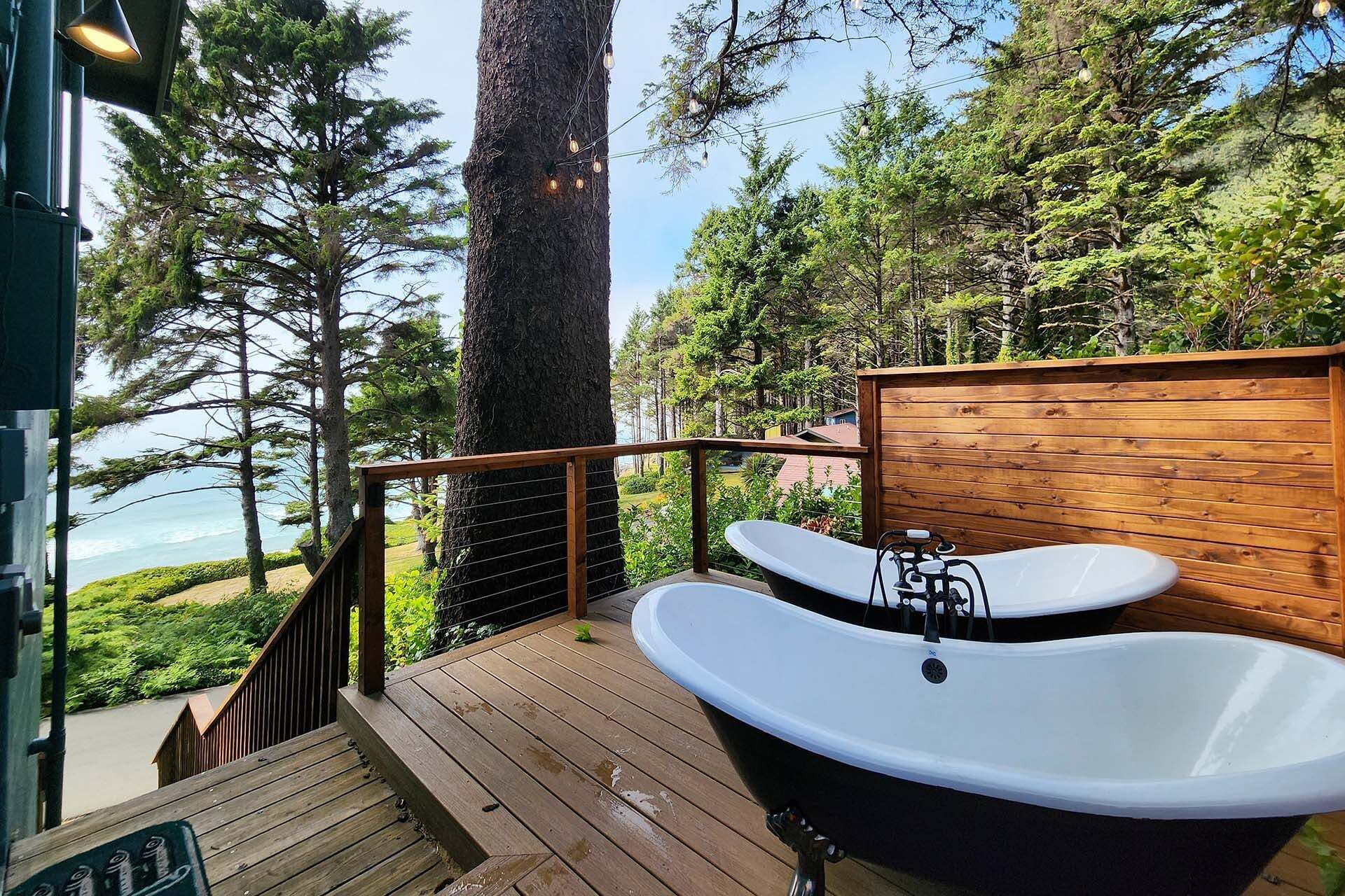 A wooden deck with a black bathtub and a white sink, overlooking a forested area and a body of water.