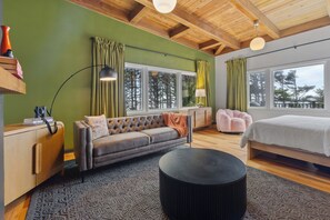 Un dormitorio moderno con una cama grande, un sofá y una mesa redonda. Hay ventanas con cortinas, techo de madera y vistas a árboles.