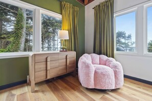 Una habitación con un armario de madera, un sillón informal rosa y dos ventanas con cortinas.