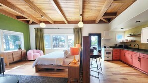 A modern bedroom with a large bed, a sitting area with a chair, a dining table, and a kitchen with red cabinets. - Ocean Front, The Pink Oyster @ Betty's Beachside, Stairs to the Beach! (Yachats)