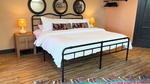 Una cama con estructura de metal negro, ropa de cama blanca y almohadas coloridas. Hay dos mesitas de noche con lámparas, suelo de madera y una alfombra estampada.