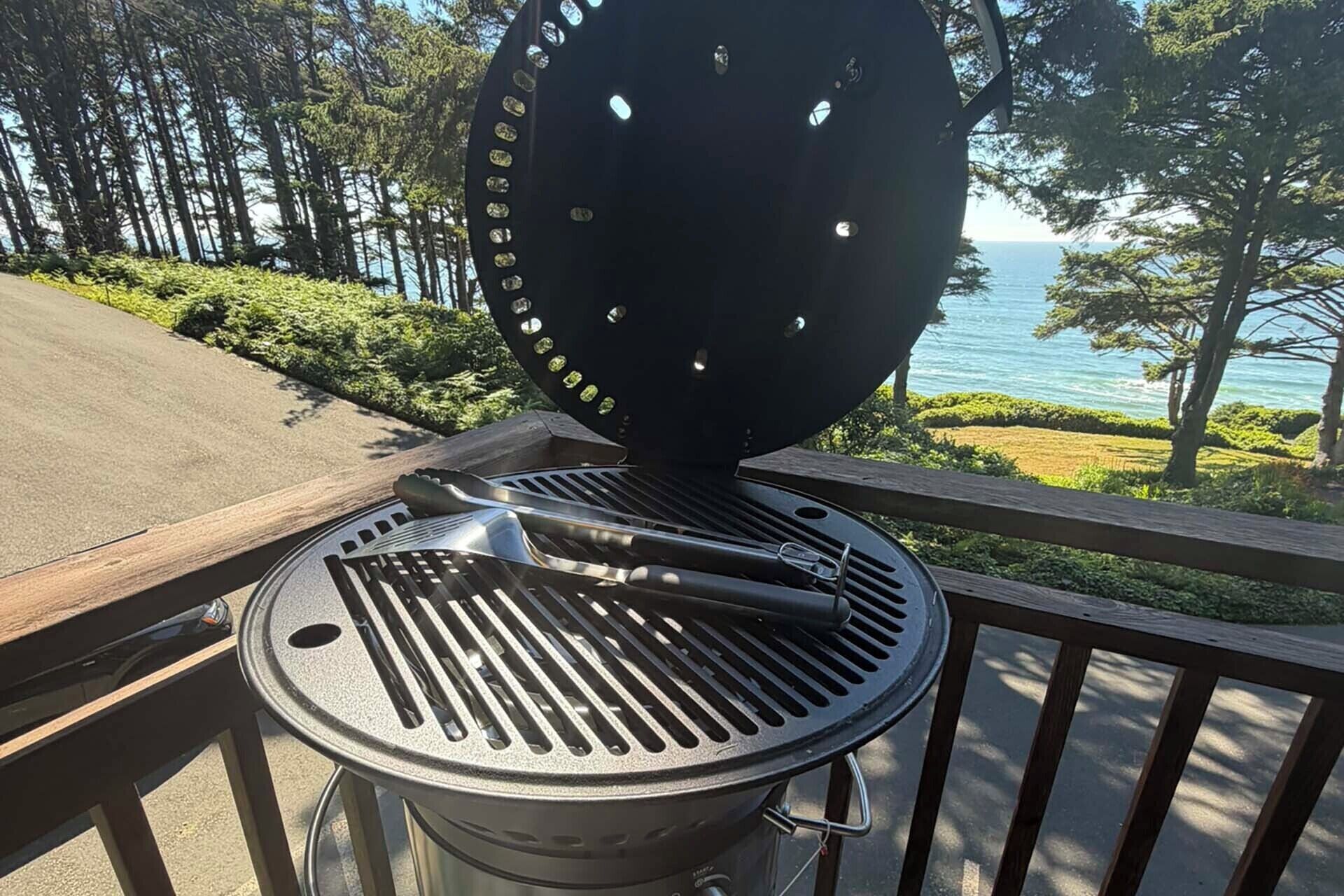 Un barbecue avec pinces et couvercle, placé sur un balcon donnant sur une route et un plan d’eau.