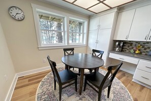 Un comedor con una mesa redonda y seis sillas, una ventana con vista a árboles, y una cocina con armarios blancos y una cafetera.